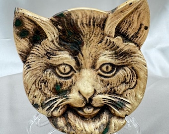 May include: A vintage-style cat face sculpture with a detailed, textured surface. The sculpture features a cream and brown color palette with green accents. The cat's face is round with pointed ears, and it is mounted on a clear stand.