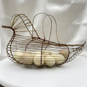 May include: A vintage-style wire bird-shaped basket, holding several speckled eggs. The basket is made of rusty brown metal wire, with a long beak and curved handle. The eggs are off-white with brown speckles, creating a rustic farmhouse aesthetic.