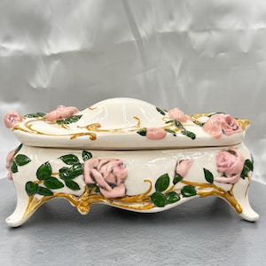 May include: A white ceramic jewelry box with a lid, adorned with pink roses and green leaves. The box has a curved shape and rests on four curved feet. Gold accents highlight the floral design.