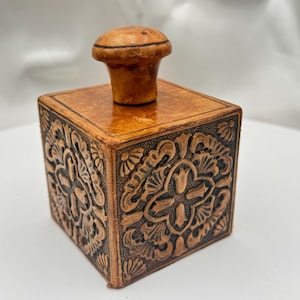 May include: A square, light brown leather box with a decorative floral pattern embossed on all sides. The box has a matching light brown, mushroom-shaped lid. The box is a decorative storage item.