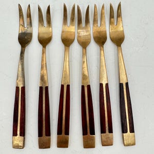 May include: Set of six vintage serving forks. The forks have a brass finish with a three-pronged design. The handles are a dark wood color with brass accents. The forks are arranged in a row.