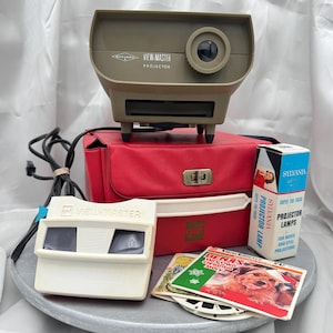 May include: A tan Sawyer's View-Master projector, a red carrying case with a gaf logo, a white View-Master viewer, a Sylvania projector lamp box, and a Benji's Very Own Christmas Story View-Master reel packet with a black power cord.
