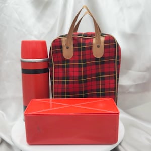 May include: A vintage picnic set featuring a red thermos, a red rectangular container, and a plaid picnic bag with brown leather handles. The bag has a red, black, and yellow plaid pattern.