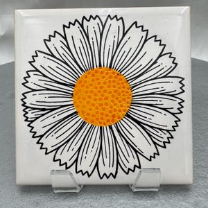 May include: Square white ceramic tile featuring a large daisy design. The daisy has a bright orange center and black and white petals. The tile is displayed on a clear plastic stand.
