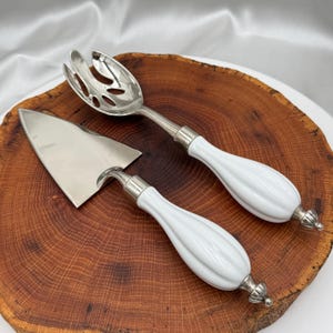 May include: A silver salad serving spoon and pie server with white ceramic handles. The serving utensils are displayed on a round, wooden cutting board with a natural wood grain pattern.