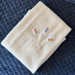 May include: A cream-colored textured blanket featuring an embroidered design of three balloon-shaped motifs. Each balloon contains a small character illustration with the names Flopsy, Cotton-Tail, and Mopsy. A stitched bow ties the balloon strings.