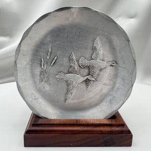 May include: A pewter-colored decorative plate with a raised relief of two flying ducks and cattails. The plate has a scalloped edge and is mounted on a dark brown wooden base. The plate is round.