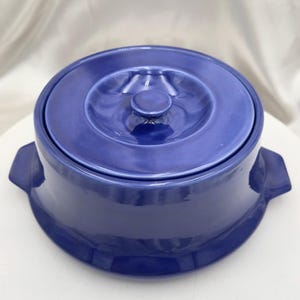 May include: A cobalt blue ceramic casserole dish with a lid. The dish has two handles and a round lid with a small knob. The dish is on a white surface.