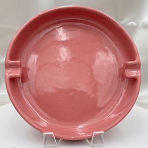 May include: A round, pink ceramic ashtray with two built-in cigarette rests. The ashtray has a smooth, glossy finish and is supported by a clear stand. The color is a muted, dusty rose.