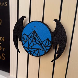 Peut inclure: Un serre-livres noir avec un cercle bleu représentant une chaîne de montagnes et des étoiles. Le serre-livres est en forme d'ailes. Le serre-livres est sur une étagère avec d'autres livres.