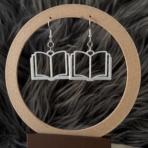Puede incluir: Pendientes plateados con forma de libros abiertos. Los pendientes cuelgan de ganchos plateados y se exhiben sobre un fondo neutro. Estos pendientes de temática literaria son un accesorio único.