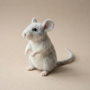 Puede incluir: Una figura de ratón de fieltro gris claro, hecha a mano, con vientre blanco, patas rosas y una larga cola. El ratón está en posición vertical, con los brazos ligeramente levantados. El fondo es beige neutro.