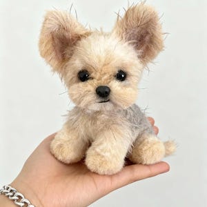 May include: A plush Yorkshire Terrier dog with tan and gray fur, large ears, and black eyes. The handmade stuffed animal is sitting upright, showcasing its detailed features and realistic appearance. The toy is held in a hand.