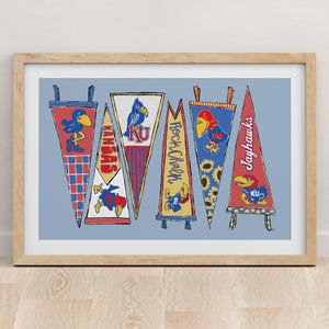 Op de afbeelding: Een ingelijste kunstdruk met zes driehoekige wimpels in rood, blauw en geel. De ontwerpen bevatten de tekst Kansas, Rock Chalk en Jayhawks, naast illustraties van een blauwe vogelmascotte, zonnebloemen en geblokte patronen.