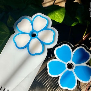 May include: Two flower-shaped accessories with a blue and white design. One is a bookmark with a comb-like edge, and the other is a pin. The accessories have a floral design with a blue outline and a white center. The background includes green leaves.