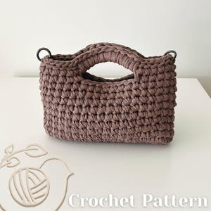 May include: A mauve-colored handbag crafted with a thick, textured crochet stitch. The bag features two integrated handles and two dark metal rings attached to the top corners. The text "Crochet Pattern" is visible in the bottom right corner.