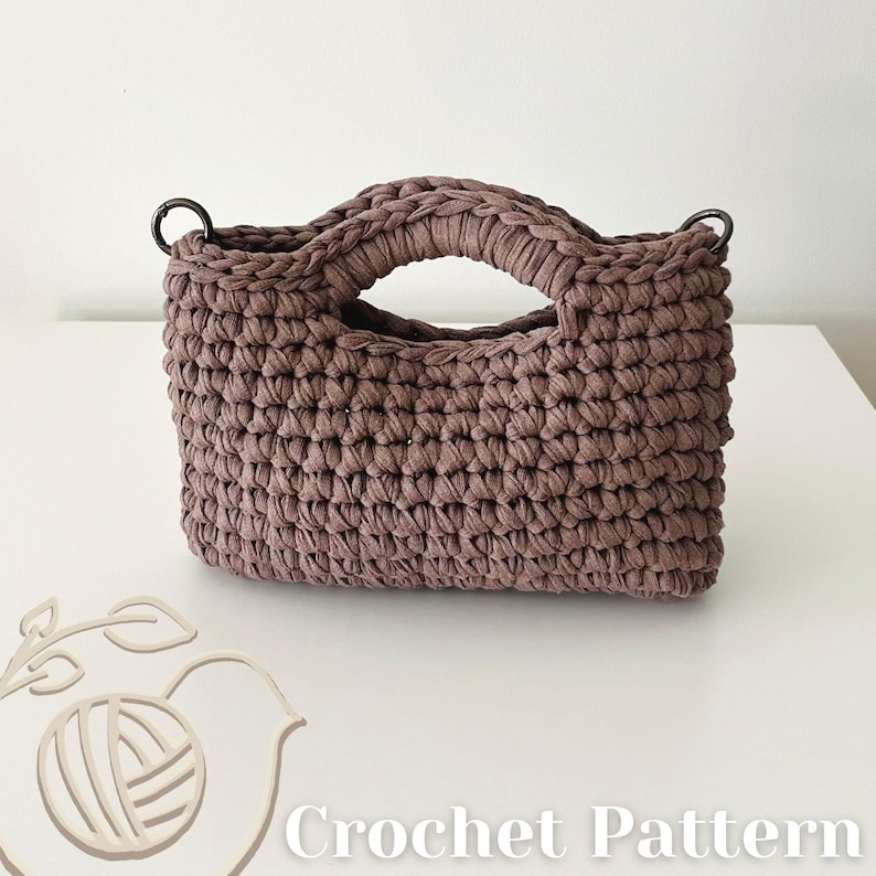 May include: A mauve-colored handbag crafted with a thick, textured crochet stitch. The bag features two integrated handles and two dark metal rings attached to the top corners. The text "Crochet Pattern" is visible in the bottom right corner.