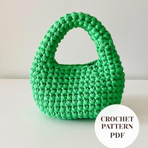 Puede incluir: Un bolso de mano de color verde brillante elaborado con un punto de ganchillo grueso y texturizado. El bolso presenta una forma redondeada de media luna con un asa integrada. Una insignia blanca circular en la esquina inferior derecha muestra el texto CROCHET PATTERN PDF.
