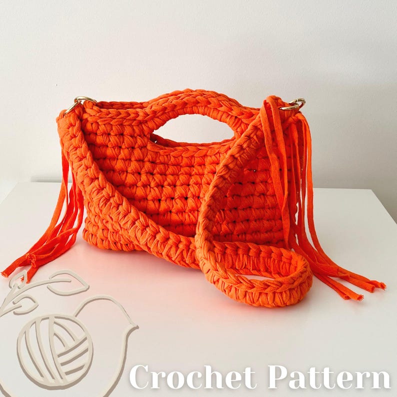 May include: A bright orange crochet handbag featuring a thick braided texture, integrated top handles, and long fringe detailing on both sides. The bag includes a matching crochet shoulder strap and gold-toned metal hardware rings.