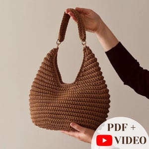 Könnte beinhalten: Eine braune Häkel-Hobo-Tasche mit strukturiertem Rippenmuster, einem einzelnen integrierten Griff und goldfarbenen Metallringen, die den Griff mit dem Körper verbinden. Das Bild enthält den Text PDF + VIDEO mit einem roten YouTube-Play-Symbol.