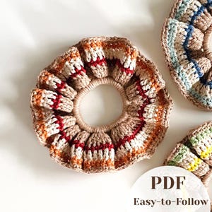 May include: A circular crochet hair scrunchie featuring a tan base with horizontal stripes of white, orange, and deep red. The design includes a ruffled texture and a central elastic opening. A white circular overlay displays the text PDF Easy-to-Follow.