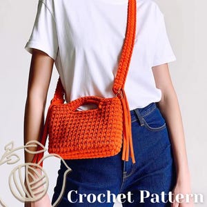 May include: A bright orange crochet handbag featuring a rectangular body with a top handle and a long shoulder strap. The bag is crafted with a thick, textured yarn stitch and includes decorative fringe accents on the sides. The text Crochet Pattern is visible.
