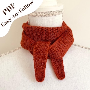 Puede incluir: Una bufanda de ganchillo de color óxido se muestra en un maniquí. La bufanda está hecha de un hilo grueso y tiene un aspecto texturizado. El texto "PDF Easy-to-Follow" está en la esquina superior izquierda. La bufanda está anudada.