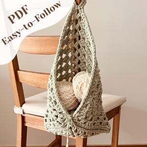 May include: A sage green crochet hanging basket with two balls of yarn inside, suspended from a wooden chair. The basket has a textured, open weave design. Text on the upper left reads "PDF Easy-to-Follow".