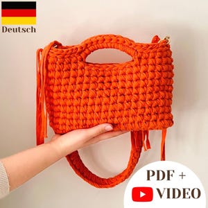 May include: A bright orange crocheted handbag featuring a rectangular body with a cutout handle at the top, a thick braided shoulder strap, and long fringe tassels hanging from the sides. The texture is consistent with thick yarn.