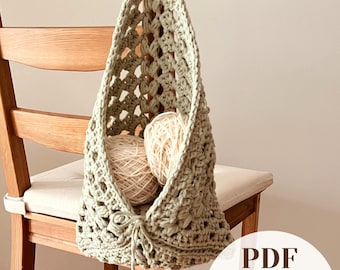 May include: A sage green crocheted hanging basket with an openwork pattern, featuring a drawstring closure at the bottom and containing two balls of cream-colored yarn. The image includes the text PDF Easy-to-Follow.
