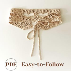 以下が含まれることがあります： ベージュのかぎ針編みトップス。ハート型のカットアウトデザインとリボン結び。左下隅に「PDF Easy-to-Follow」の文字。柔らかく、質感のある糸で作られています。