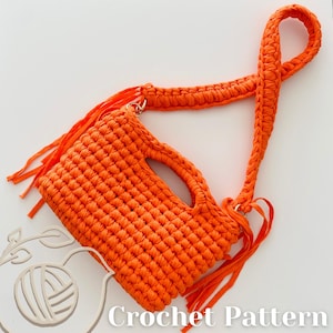 May include: A bright orange crochet handbag featuring a thick braided shoulder strap, a rectangular body with a cutout handle, and long decorative fringe accents on the sides. The bag is constructed from a chunky yarn with a visible stitch pattern.
