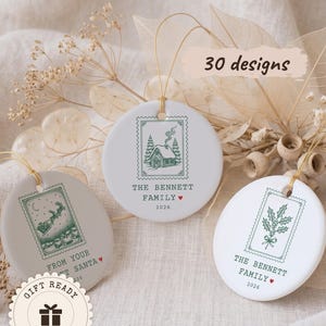 May include: Three round white and light gray ceramic ornaments with gold hanging strings. Each features a green postage stamp illustration: a cabin, a sleigh, or holly. Text includes "THE BENNETT FAMILY", "2026", "FROM YOUR SANTA", and "30 designs".