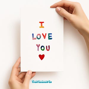 Puede incluir: Una tarjeta de felicitación blanca con las palabras "I LOVE YOU" en letras coloridas, estilo acuarela. Un corazón rojo está debajo del texto. La tarjeta está sostenida por dos manos.