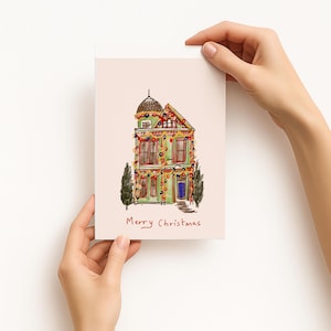 May include: A rectangular greeting card with a light pink background featuring a hand-drawn illustration of a green house decorated with colorful Christmas lights. The text Merry Christmas is written in red ink below the house illustration.