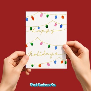 May include: A white greeting card featuring gold string lights with colorful painted bulbs in blue, green, pink, and orange. The text "happy holidays" is written in gold cursive script across the center of the card between the light strings.