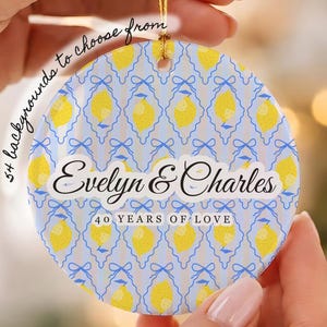 Puede incluir: Un adorno redondo de cerámica con un patrón repetitivo de limones amarillos y lazos azules sobre un fondo de rayas azul claro y blanco. El texto dice Evelyn & Charles y 40 YEARS OF LOVE. Un cordón dorado está sujeto en la parte superior.