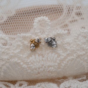 May include: Two stud earrings, one gold-toned and one silver-toned, each featuring a cluster of clear, sparkling cubic zirconia stones. The earrings are displayed on a white lace surface, creating a delicate and elegant presentation.