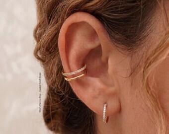 Peut inclure: Gros plan sur une oreille ornée de bijoux en or. L'oreille est ornée d'un double ear cuff avec des pierres scintillantes et d'une petite boucle d'oreille avec des pierres similaires. Les bijoux se détachent sur la peau et les cheveux de la personne.