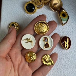 Puede incluir: Una colección de botones decorativos variados en diferentes formas y acabados, incluyendo metal dorado, esmalte blanco con motivos dorados, un diseño de ojo en blanco y negro y una pieza de color rojo oscuro, sobre una superficie blanca texturizada.