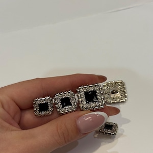 May include: Several square, silver-coloured buttons with black centres and rhinestone borders. The buttons are of varying sizes and are shown on a white surface. These buttons are likely for clothing or craft projects.
