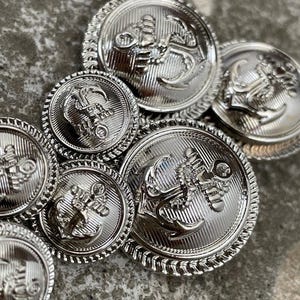 May include: A collection of shiny silver metal buttons with an anchor design. The buttons vary in size and have a detailed rope-like border. The anchor is centered on each button, with intricate detailing. These buttons are likely for clothing or craft projects.