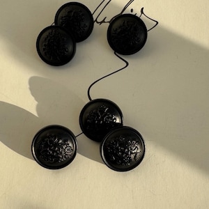 Può includere: Sei bottoni neri con intricati motivi floreali disposti su una superficie chiara. I bottoni sono circolari e presentano un disegno in rilievo. Un'ombra è proiettata sulla scena. I bottoni sembrano essere fatti di un materiale duro.