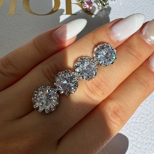 May include: Four round, silver-toned stud earrings with clear, sparkling gemstones are displayed. The earrings are arranged in a row, showcasing their intricate design and brilliance. A second, smaller earring with colorful gemstones is also visible.