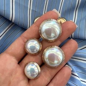 Peut inclure: Quatre boutons nacrés avec des bords en métal doré. Les boutons sont de tailles différentes et ont une finition légèrement irisée.