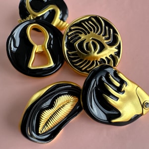 May include: A collection of black and gold-colored decorative buttons. The buttons feature various designs, including a keyhole, an eye, and abstract shapes. The buttons have a glossy finish and are arranged on a pink surface.