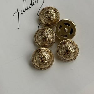 May include: A collection of gold-colored buttons with a detailed Medusa head design. The buttons have a raised, textured border and a shiny finish. One button shows the back with a loop for sewing. The background is a white surface with a handwritten signature.