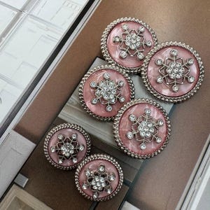 May include: Six decorative buttons with a pink marbled centre and a silver-coloured rope-like border. Each button features a snowflake design with clear crystal-like accents. The buttons are arranged on a magazine.