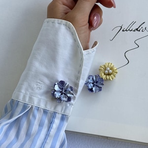 May include: A close-up of a white and blue striped shirt cuff with a blue flower cufflink. Two additional flower cufflinks, one blue and one gold, are displayed on a white surface. The gold cufflink has three small pearls in the center.