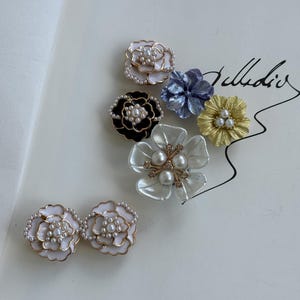 Peut inclure: Un assortiment de bijoux en forme de fleurs, comprenant des boucles d'oreilles et des broches. Les pièces présentent des pétales blancs, noirs, dorés et bleus, avec des perles. Les bijoux sont disposés sur une surface blanche, mettant en valeur les détails et les motifs complexes.